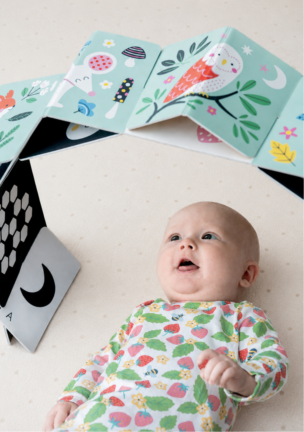 A baby lying on a light beige floor, looking up at a cardboard arch illustrated with colourful animals on the outside, and high-contrast black and white patterns and shapes on the inside. The baby is wearing a white long-sleeve onesie with a strawberry and leaf print.