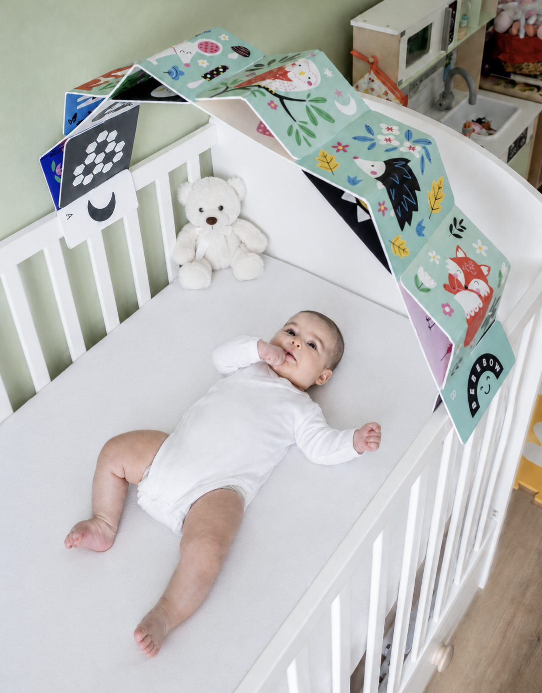 Baby lying in a crib with baby sensory arch and a teddy bear